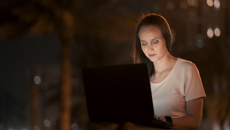 A-young-female-student-with-a-laptop-at-night-in-the-city-looking-at-the-computer-screen-and-typing-with-his-hands-on-the-keyboard.-Remote-work-on-the-Internet.-The-student-does-the-work.-Night-life-in-the-city